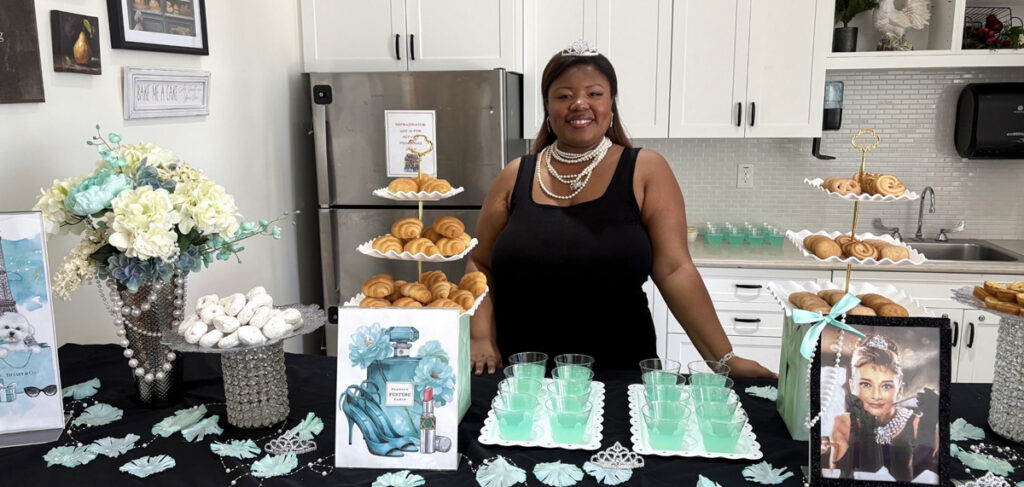 An employee standing behind a decorated refreshment table at a senior living event, featuring pastries, teal drinks, flowers, and themed decor with an Audrey Hepburn image in a bright kitchen setting.