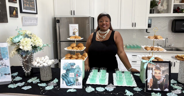 An employee standing behind a decorated refreshment table at a senior living event, featuring pastries, teal drinks, flowers, and themed decor with an Audrey Hepburn image in a bright kitchen setting.