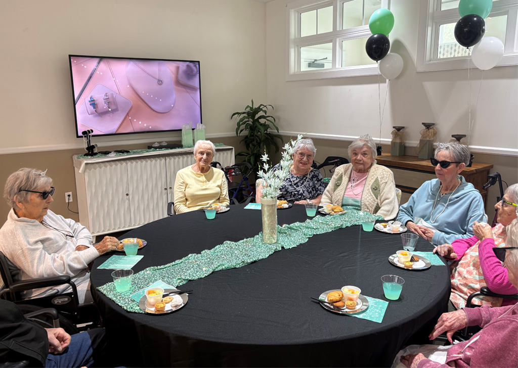 Residents gathered around a round table at a senior living community, wearing sunglasses and enjoying refreshments during a themed activity, with black, white, and green balloons and a TV screen in the background.