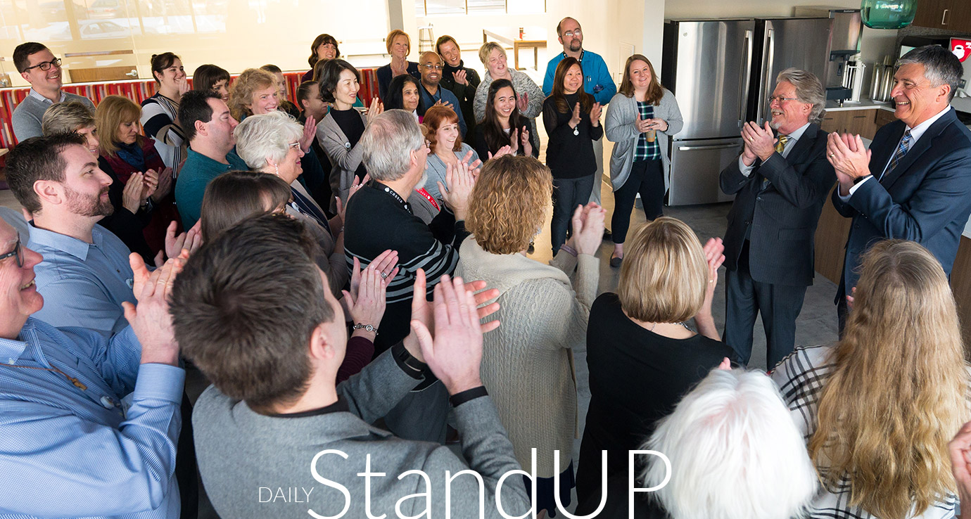 Large group of Cassia team members gathered in a common area during a daily standup meeting, with people applauding and the text “Daily StandUP” displayed