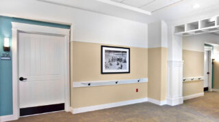 Apartment hallway with resident entry door, decorative wall art, handrails, wood trim, and wide accessible corridors at Trinity Springs