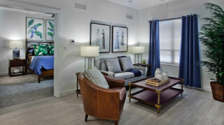 Apartment living room with sofa, armchair, coffee table, large window, and adjoining bedroom in a spacious residential floor plan at Trinity Springs