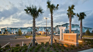 Exterior view of Trinity Springs featuring the community entrance, landscaped grounds, palm trees, parking area, and illuminated monument sign at dusk