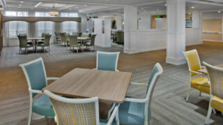 Open community gathering space with tables, upholstered chairs, wide walkways, large windows, and flexible seating areas for games, activities, and social events at Trinity Springs
