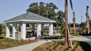 Covered outdoor lanai with seating, walking paths, palm trees, landscaped gardens, and fenced courtyard space at Trinity Springs