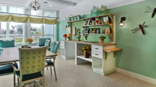 Craft and activity room with tables, chairs, workstations, decorative shelving, floral accents, and large windows providing natural light at Trinity Springs