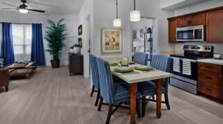 Spacious apartment with a full kitchen, dining table, stainless steel appliances, living room seating, large windows, and open floor plan at Trinity Springs