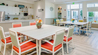 Memory care dining area with tables, chairs, community kitchen, large windows, and direct access to an outdoor courtyard at Trinity Springs