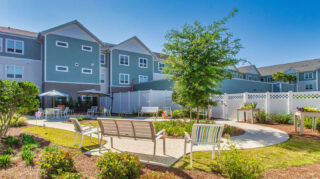 Memory care garden courtyard with walking paths, benches, patio seating, umbrellas, landscaped flower beds, and outdoor gathering areas at Trinity Springs