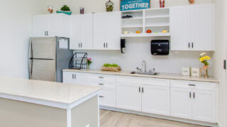 Memory care kitchen with white cabinetry, center island, stainless steel refrigerator, sink, countertop workspace, and open shelving at Trinity Springs