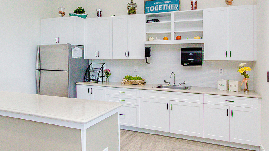 Memory care kitchen with white cabinetry, stainless steel refrigerator, sink, countertop workspace, and center island in a bright community setting at Trinity Springs