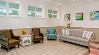 Memory care lounge with sofa seating, upholstered chairs, side table, artwork, and comfortable gathering space at Trinity Springs