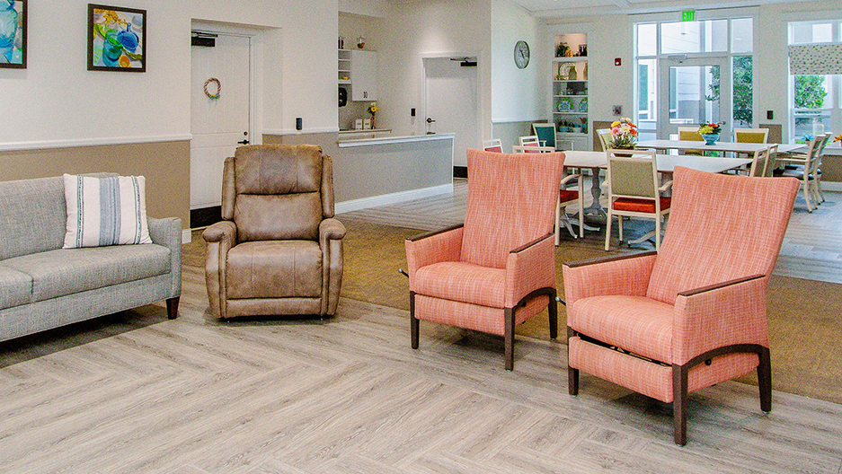 Memory care lounge with comfortable seating, dining tables, natural light, and an open gathering space connected to the community kitchen at Trinity Springs