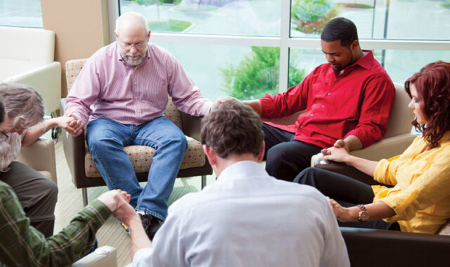 Group of adults seated in a circle holding hands in a shared gathering space near large windows, representing faith-based activities and spiritual support like at Trinity Springs